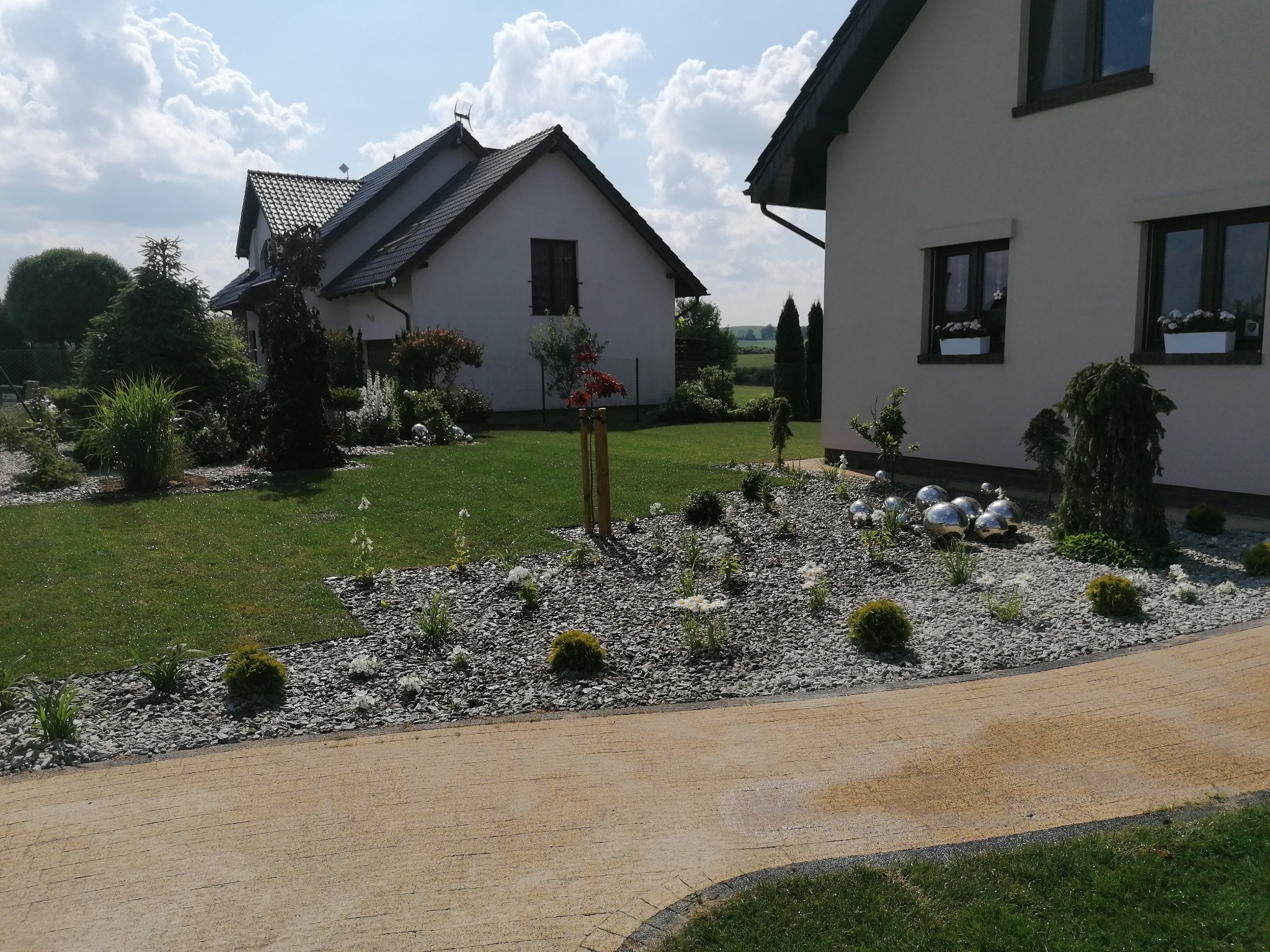 Uporządkowany ogród z trawnikiem, ścieżką z kostki brukowej i rabatami wysypanymi szarym żwirem, ozdobiony srebrnymi kulami, na tle dwóch domów jednorodzinnych w słoneczny dzień.