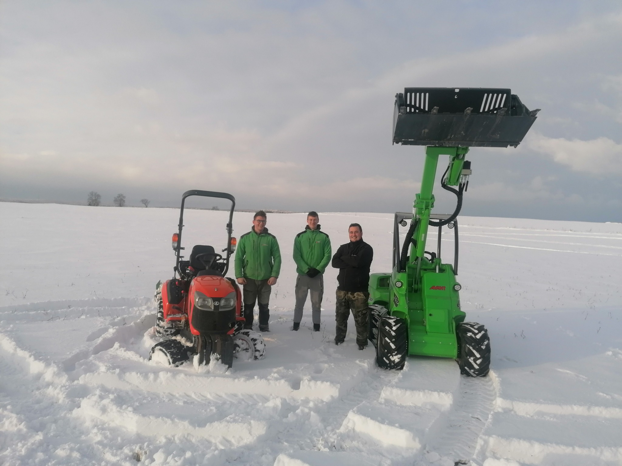 Trzech mężczyzn w zimowej scenerii stoi między małym czerwonym traktorem a zieloną ładowarką teleskopową na tle zaśnieżonego pola i pochmurnego nieba.