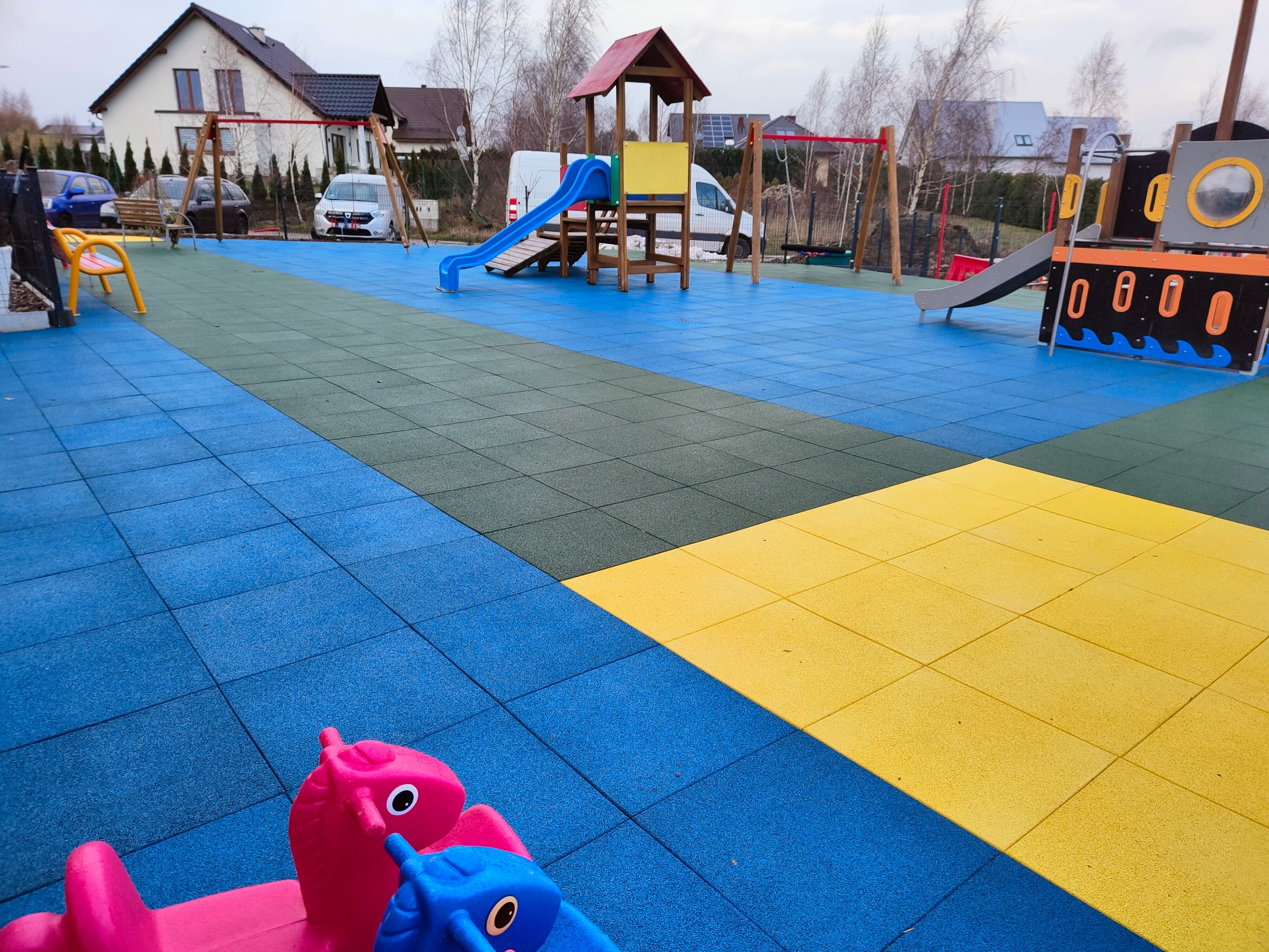 Kolorowa nawierzchnia placu zabaw z gumowych płyt w odcieniach niebieskiego, zielonego i żółtego, z widocznymi urządzeniami zabawowymi: zjeżdżalnie, huśtawki i bujaki w kształcie zwierząt.
