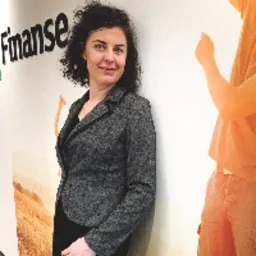 Uśmiechnięta kobieta w szarej marynarce, oparta o ścianę z napisem 'Finanse' i częściowo widoczną grafiką osoby.