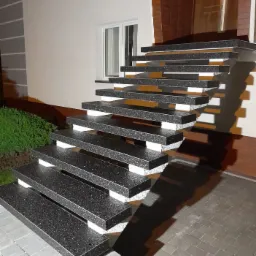 Nowoczesne, zewnętrzne schody z czarnego kamienia z białymi podstopnicami, prowadzące do drzwi wejściowych budynku o minimalistycznej architekturze, oświetlone ciepłym światłem.