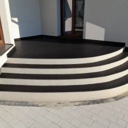 Czarny taras z żywicy z czterema zaokrąglonymi schodami, wykończony jasnoszarą kostką brukową i oknem w tle.