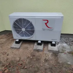 Biała pompa ciepła Rotenso Aquami Series zamontowana na betonowych podstawach przy ścianie budynku o żółtej elewacji, widoczne liście i ziemia.