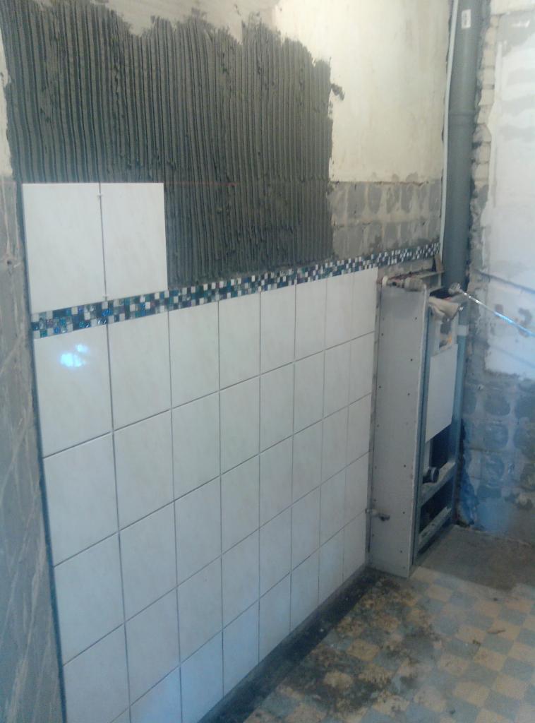 Układanie białych płytek na ścianie z dekoracyjnym pasem mozaiki. Widoczny fragment ściany z klejem i instalacja sanitarna. Prace wykończeniowe w łazience.