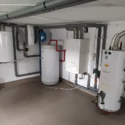 Kompleksowa instalacja grzewcza w kotłowni, widoczne dwa bojlery, piec gazowy, zbiornik wyrównawczy i rozbudowany system rur.