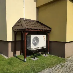 Biała jednostka zewnętrzna klimatyzatora pod drewnianą zadaszoną konstrukcją na trawniku przy budynku z elewacją w odcieniach beżu i brązu, widoczny fragment chodnika z kostki brukowej.