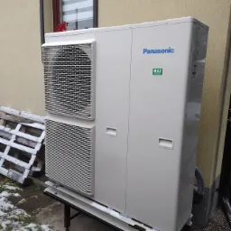 Zewnętrzna jednostka pompy ciepła Panasonic R32 zamontowana na metalowym stelażu, częściowo ośnieżona, na tle elewacji budynku z oknem.