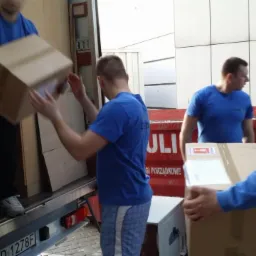 Ekipa w niebieskich koszulkach wyładowuje kartony z ciężarówki. Widoczny numer rejestracyjny pojazdu. Praca w toku, etap wyładunku.