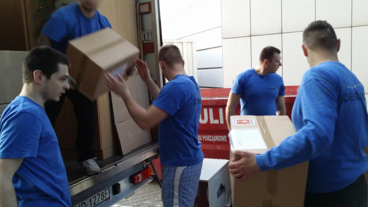 Ekipa w niebieskich koszulkach wyładowuje kartony z ciężarówki. Widoczny numer rejestracyjny pojazdu. Praca w toku, etap wyładunku.