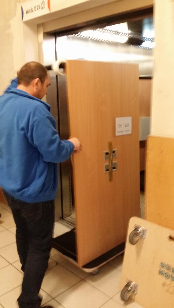 Pracownik w niebieskiej bluzie wprowadza szafę do windy za pomocą wózka transportowego na kółkach. Widoczna tabliczka 'Winda 0 21'.