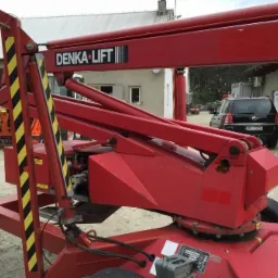 Czerwony podnośnik elektryczny DENKA LIFT DL 15 z widocznym logo, zaparkowany na zewnątrz, w tle budynek i samochód.