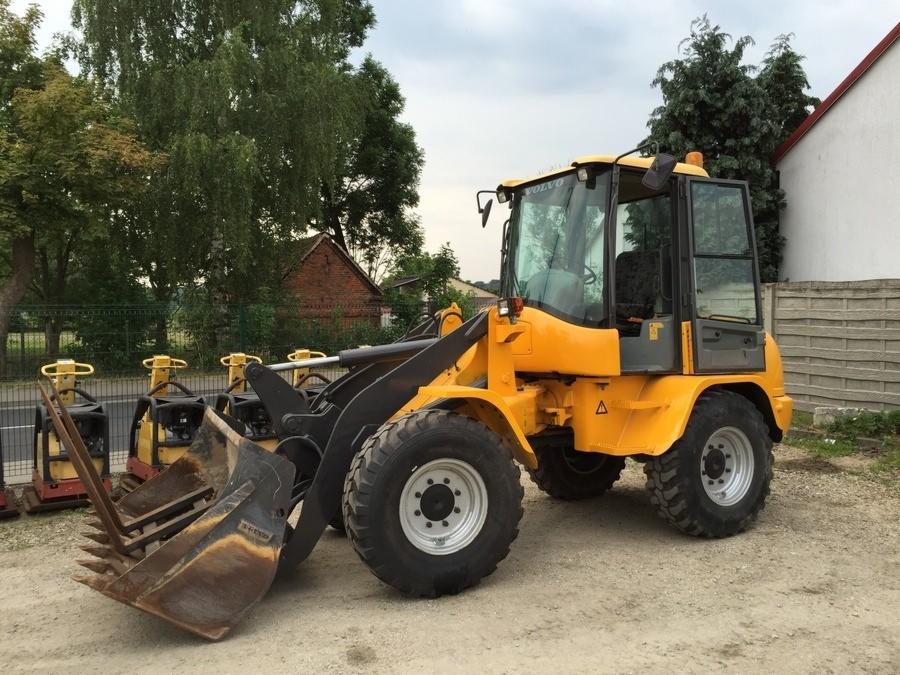 Żółta ładowarka VOLVO L 35 B-Z z łyżką i widłami na tle zieleni i budynków, obok stoją zagęszczarki.