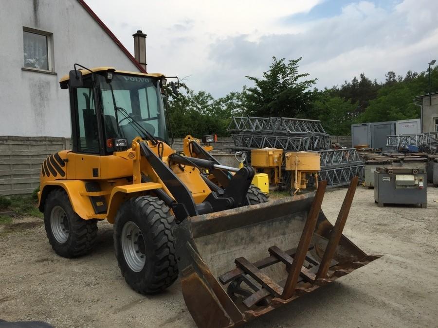 Żółta ładowarka VOLVO L 35 B-Z z widocznymi śladami użytkowania na łyżce, zaparkowana na placu budowy z betonowymi mieszalnikami i metalowymi konstrukcjami w tle.