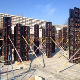 Szare szalunki kolumnowe, stabilizowane żółtymi i fioletowymi podporami, na betonowym fundamencie. W tle budynek mieszkalny i błękitne niebo.