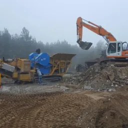 Praca ciężkiego sprzętu: koparka Liebherr, ładowarka JCB i kruszarka CAT na placu budowy w mglisty dzień. Wyburzenia i recykling materiałów budowlanych.