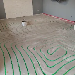 Węże systemu grzewczego w układzie meandrycznym, zalane w szlichcie podłogowej, widoczny biały pojemnik z narzędziami, ściany w odcieniach szarości i bieli.