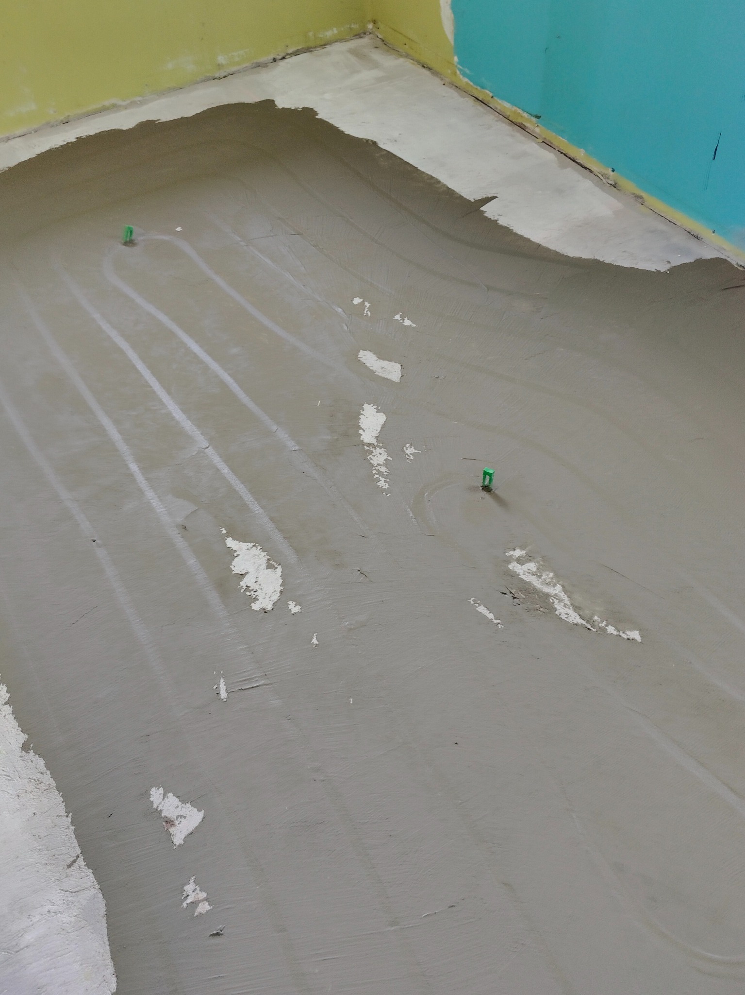 Świeżo wylany beton na rurach instalacji grzewczej, widoczne zielone uchwyty mocujące rury do podłoża, ślady po łatach na powierzchni betonu, fragment ściany w kolorze żółtym i turkusowym.
