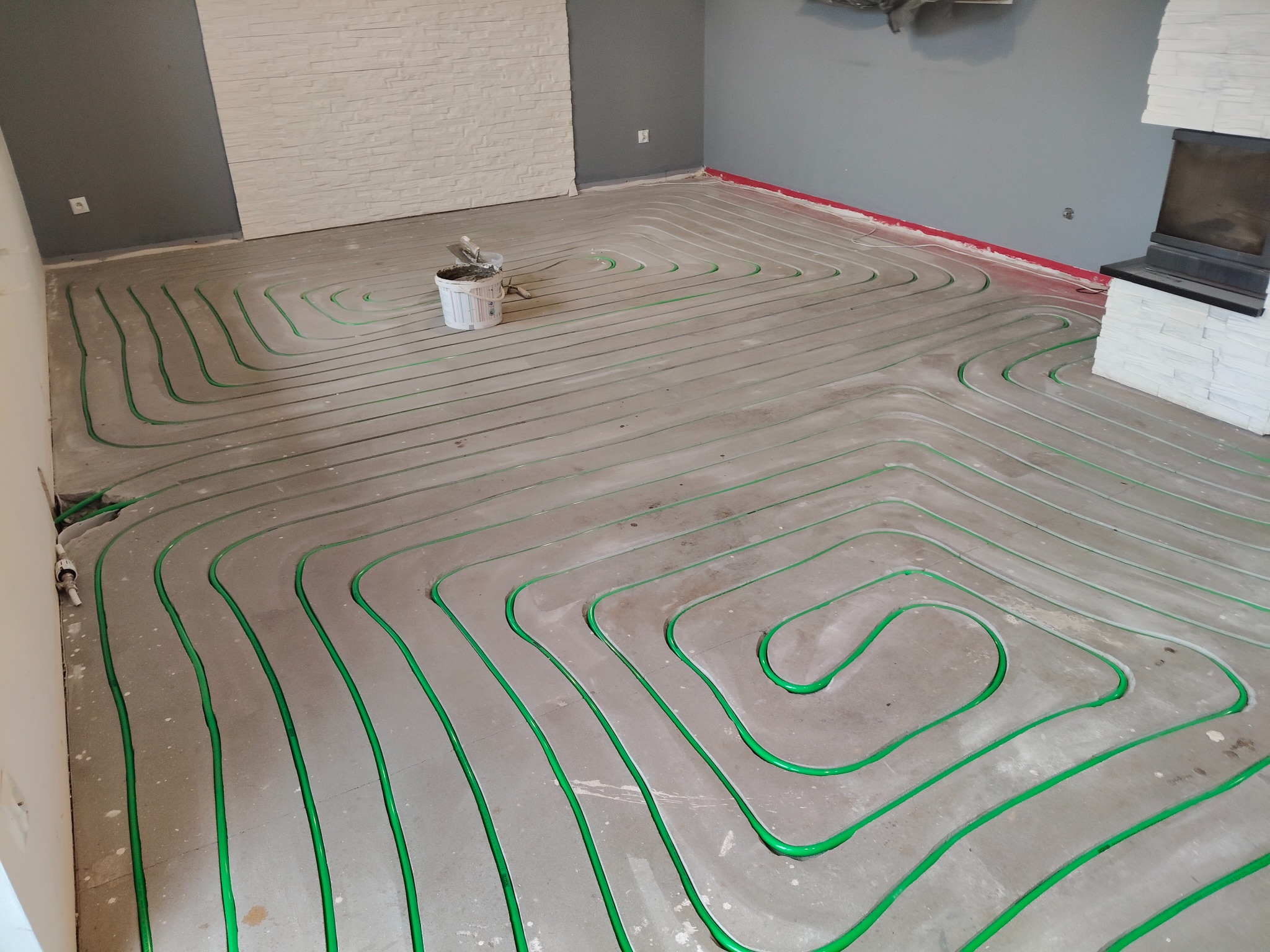 Węże systemu grzewczego w układzie meandrycznym, zalane w szlichcie podłogowej, widoczny biały pojemnik z narzędziami, ściany w odcieniach szarości i bieli.