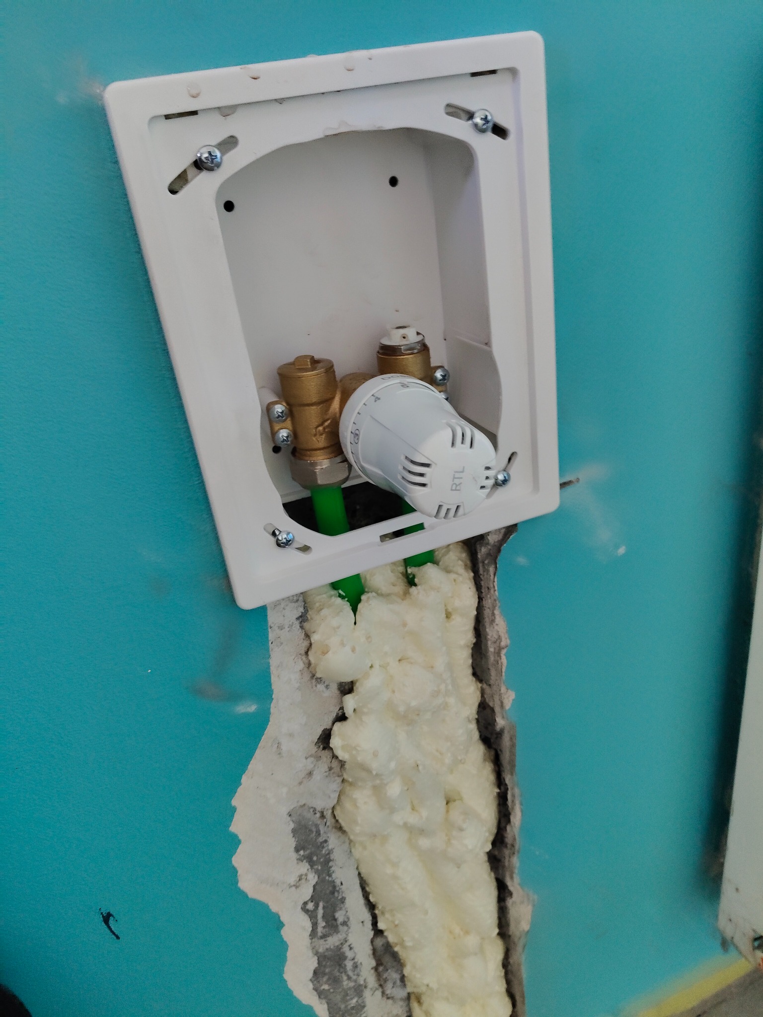Biała skrzynka montażowa z zaworami termostatycznymi RTL i zielonymi rurami, osadzona w turkusowej ścianie z widoczną pianką montażową uszczelniającą otwór.