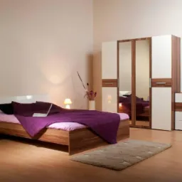Nowoczesna sypialnia z łóżkiem z drewnianą ramą, fioletową narzutą, szafką nocną z lampką, oraz szafą z lustrami i komodą w kolorze drewna i bieli.