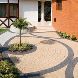 Beżowa kostka brukowa ułożona w spiralny wzór z ciemnoszarą obwódką, otaczająca małe drzewko, prowadząca do wejścia domu z kolumną i ceglaną ścianą.