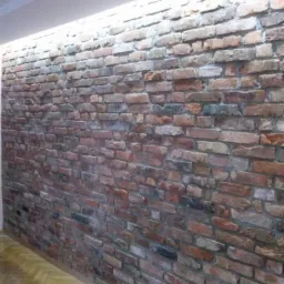 Ściana z cegły klinkierowej wewnątrz pomieszczenia z oświetleniem ledowym pod sufitem i parkietem na podłodze. Widok z dołu, ukazujący fakturę i kolorystykę cegieł.