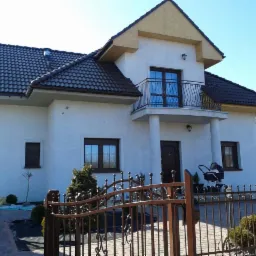 Dwukondygnacyjny dom z balkonem i brązowym dachem, widoczny zza ozdobnego, metalowego ogrodzenia. Biała elewacja, brązowe okna i drzwi. Na podjeździe wózek dziecięcy.