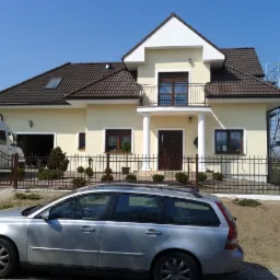 Żółty dom jednorodzinny z brązowym dachem i balkonem, ogrodzony kutym płotem. Na podjeździe stoi srebrny samochód. Widoczne rusztowanie przy ścianie.