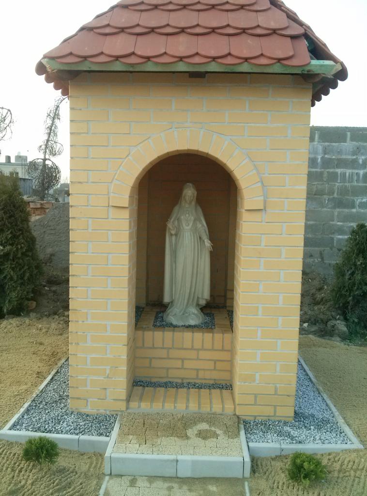 Murowana kapliczka z beżowej cegły z czerwoną dachówką, wewnątrz figura. Otoczenie wyłożone kostką i żwirem, małe krzewy. Widok z dołu na konstrukcję w ogrodzie.