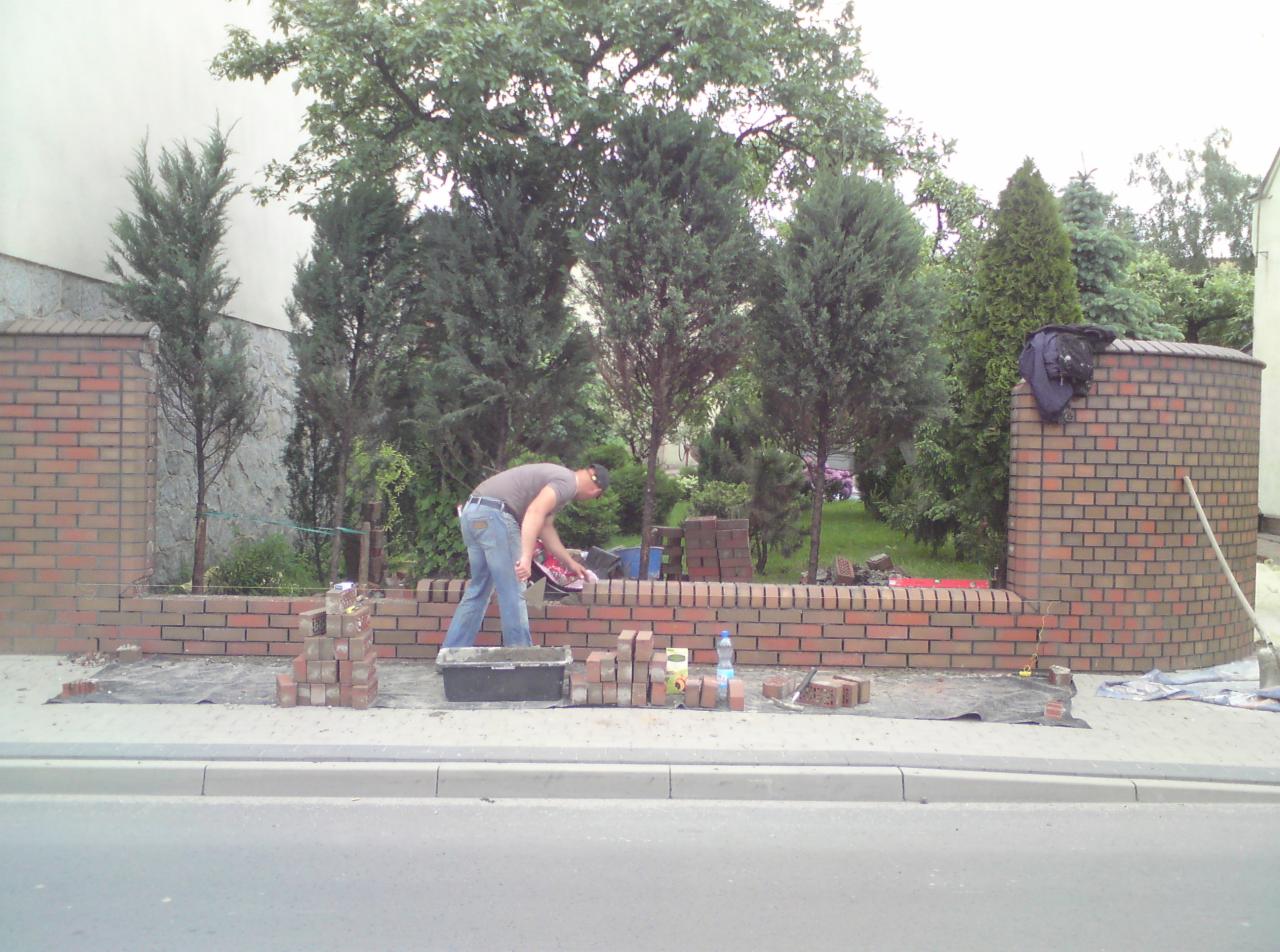 Mężczyzna w trakcie murowania niskiego murku z czerwonej cegły. Widoczne narzędzia, zapas cegieł i fragment ulicy. W tle zieleń drzew i krzewów.