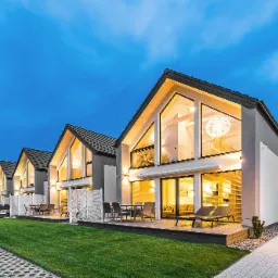 Szereg identycznych, nowoczesnych domów z dużymi oknami i drewnianymi tarasami, oświetlonych wieczornym światłem, z zadbanym trawnikiem i ścieżką.