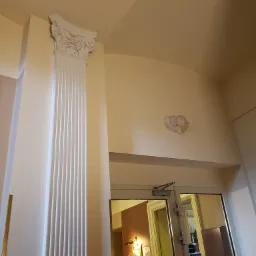 Biała kolumna dekoracyjna z ornamentem na suficie w jasnym wnętrzu, widok na lustrzane drzwi.