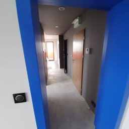 TOM-RUC Tomasz Ruczkowski - Pomalowane na biało ściany i sufit w odcieniach szarości z niebieskimi framugami drzwi i ościeżnicami, widok na korytarz z szarą podłogą i drzwiami wejściowymi.