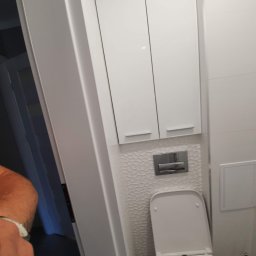 TOM-RUC Tomasz Ruczkowski - Biała toaleta z zabudową szafkową nad nią, wykończona płytkami o wzorze plastra miodu, widok z boku z fragmentem ramienia w kadrze.