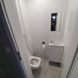 TOM-RUC Tomasz Ruczkowski - Mała, odnowiona łazienka w odcieniach bieli i szarości, z toaletą, umywalką z szafką i lustrem, widok zza uchylonych drzwi.