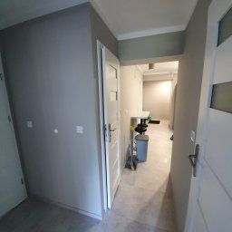 TOM-RUC Tomasz Ruczkowski - Szary korytarz po odświeżeniu, widoczne świeżo pomalowane ściany w odcieniach szarości, białe drzwi z nowoczesnymi klamkami, jasna podłoga z szarych płytek, w głębi widoczny fragment pomieszczenia...