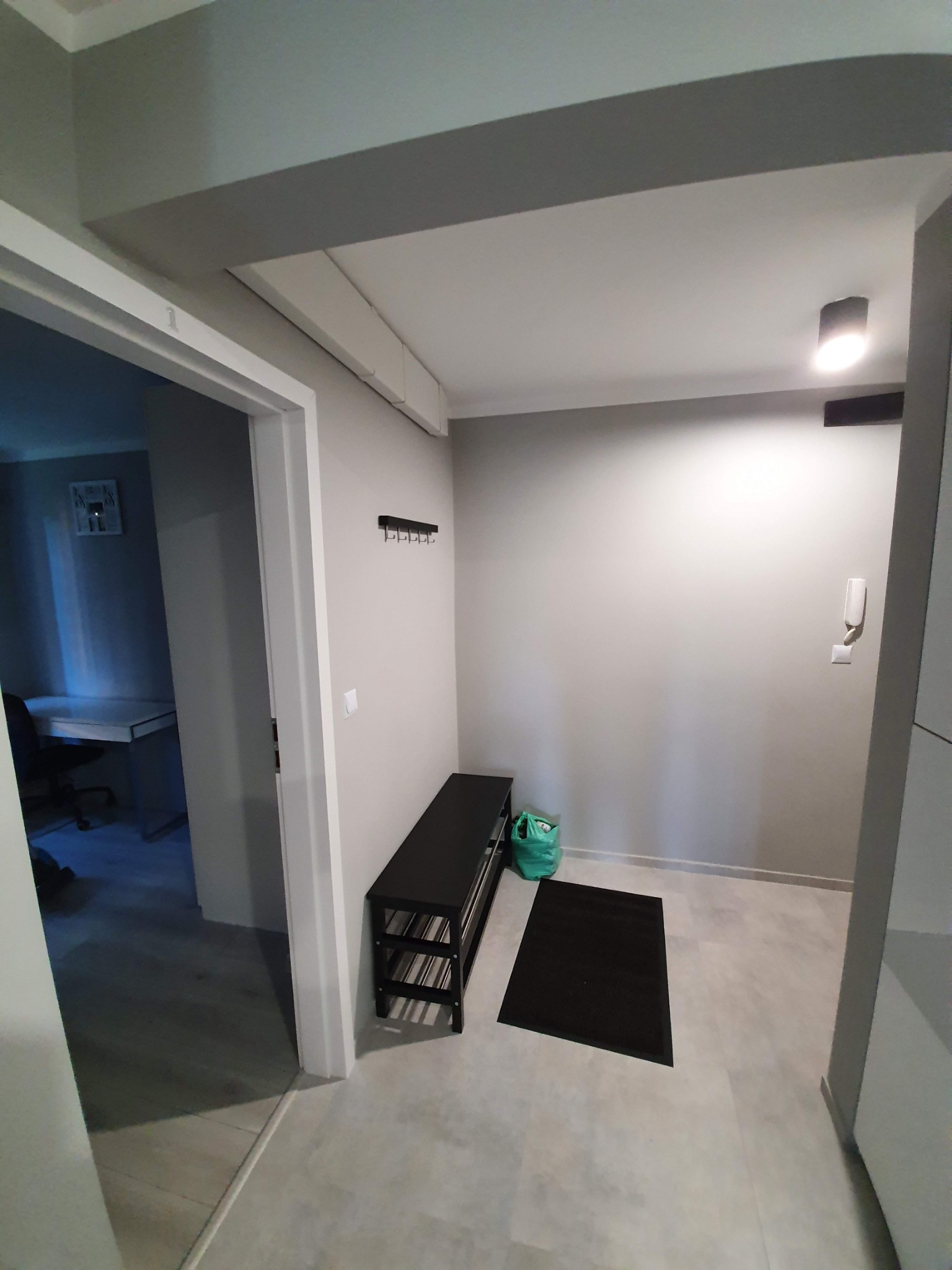 Przedpokój po odświeżeniu: szare ściany, jasna podłoga, czarna ławka na buty, czarny dywanik i wieszak na ubrania. Widoczne biuro w tle.