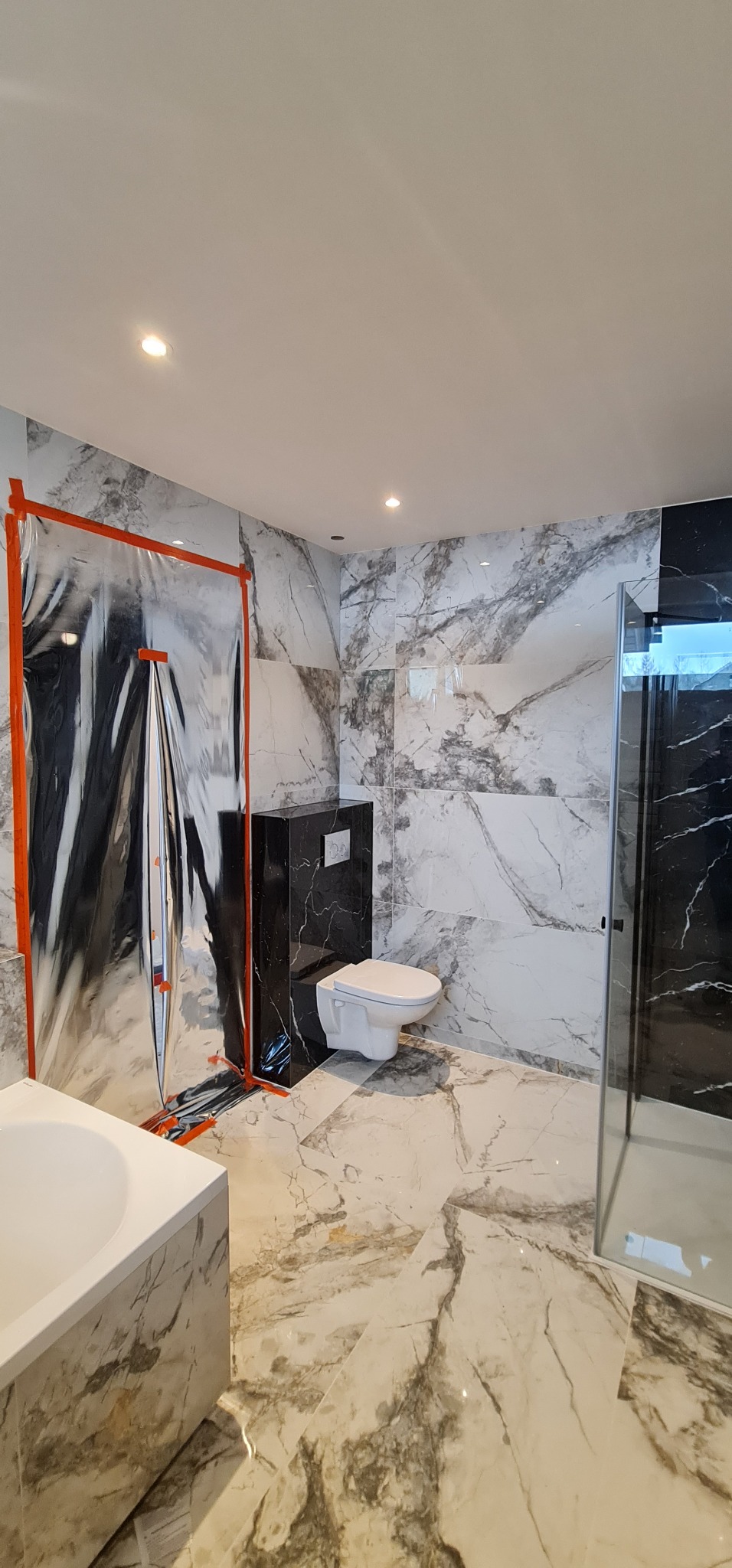 Nowoczesna łazienka w trakcie wykańczania, z marmurowymi płytkami na ścianach i podłodze, zabezpieczona folią ochronną podczas prac, widoczna toaleta podwieszana i fragment kabiny prysznicowej.