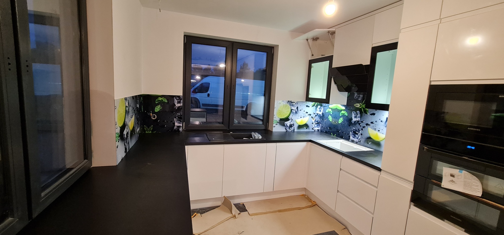Nowoczesna kuchnia z białymi szafkami i czarnym blatem, ozdobiona panelem szklanym z motywem limonki i kostek lodu, widok na okno z zaparkowanym białym vanem.