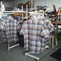Koszule w kratę na wieszakach na k&oacute;łkach w warsztacie, w tle maszyny i materiały.