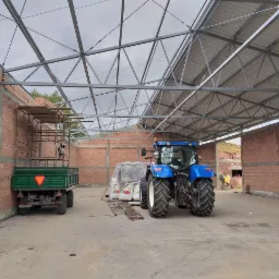 Wnętrze budowanej hali z widoczną konstrukcją stalową dachu, ścianami z czerwonej cegły, niebieskim traktorem i przyczepą rolniczą w środku. Częściowo zadaszona hala w trakcie budowy.