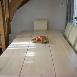 Jasny, drewniany stół z trzema krzesłami w tle, na blacie leżą jabłka w dekoracyjnej misie, obok widoczne schody.