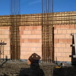 Wznoszenie ścian domu murowanego z bloczków ceramicznych, widoczna siatka zbrojeniowa, szalunek i fragment rusztowania w słoneczny dzień.