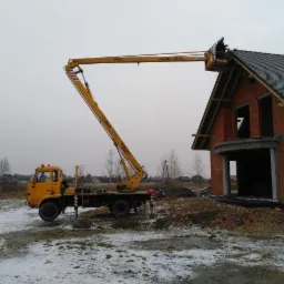 Żółty podnośnik koszowy przy murowanym domu w stanie surowym otwartym, robotnik na platformie pracuje na dachu pokrytym częściowo dachówką, na ziemi leży śnieg.