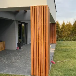 Nowoczesny taras z szarej kostki z drewnianymi pionowymi panelami, różowe kalosze przy drzwiach przesuwnych, grabie na trawie.