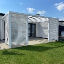 Nowoczesna pergola z białymi, poziomymi lamelami, przylegająca do jasnego budynku z dużym oknem, otoczona zielonym trawnikiem i błękitnym niebem z lekkimi obłokami.