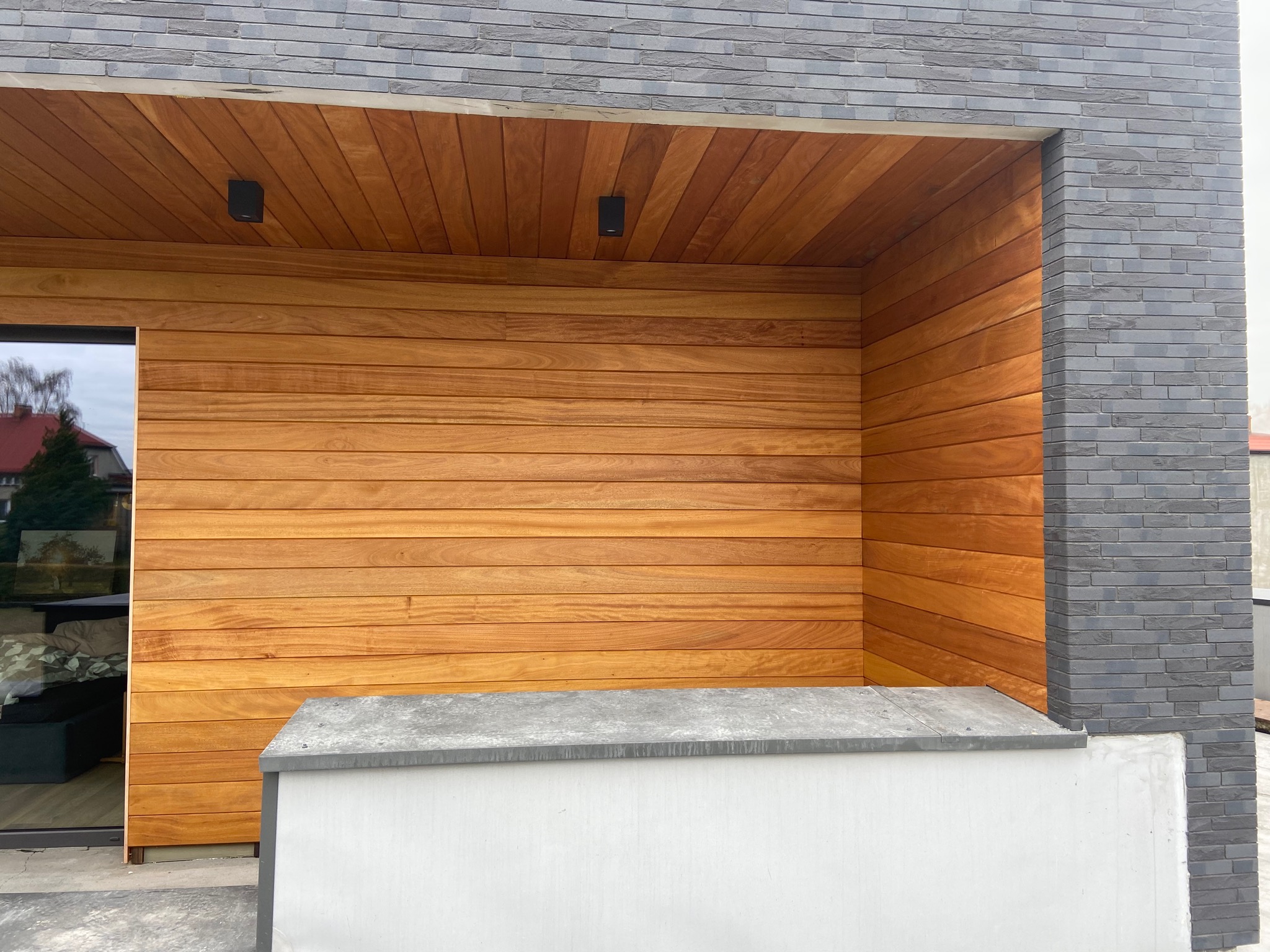 Nowoczesna elewacja z poziomych desek drewnianych i szarej cegły, z dwoma czarnymi reflektorami wbudowanymi w drewniany sufit pod okapem.