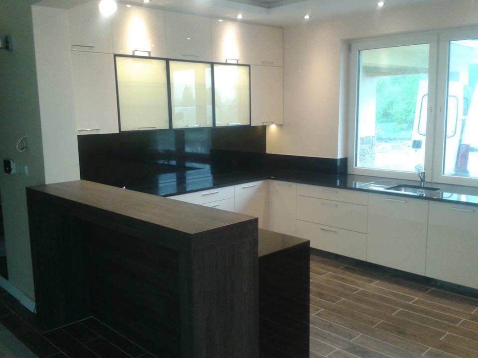 Nowoczesna, biała kuchnia z czarnym blatem i barkiem z ciemnego drewna. Uwagę zwracają podświetlane szafki górne i duże okno z widokiem na zieleń.