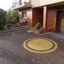 Ułożona kostka brukowa w odcieniach brązu i żółci tworzy wzór koła na podjeździe. Widoczny fragment domu z cegły i drewniana ławka z kwiatami.