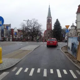 Nowa nawierzchnia drogi i chodnika z kostki brukowej w Rumi. Widoczne oznakowanie poziome, budynki w tle i czerwony samochód na jezdni.